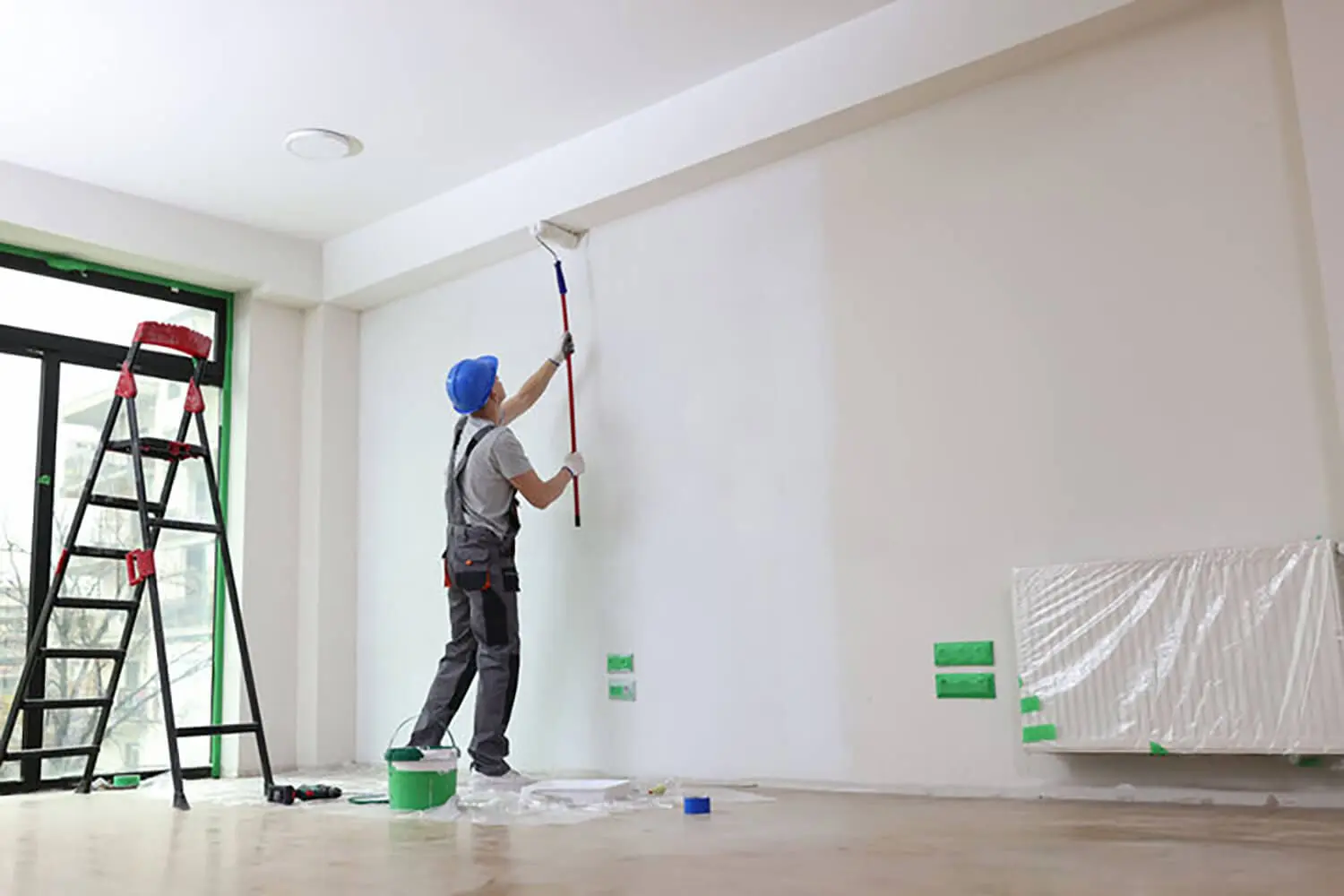 Professional painter rolling white paint on an interior wall with drop cloths and ladder in place
