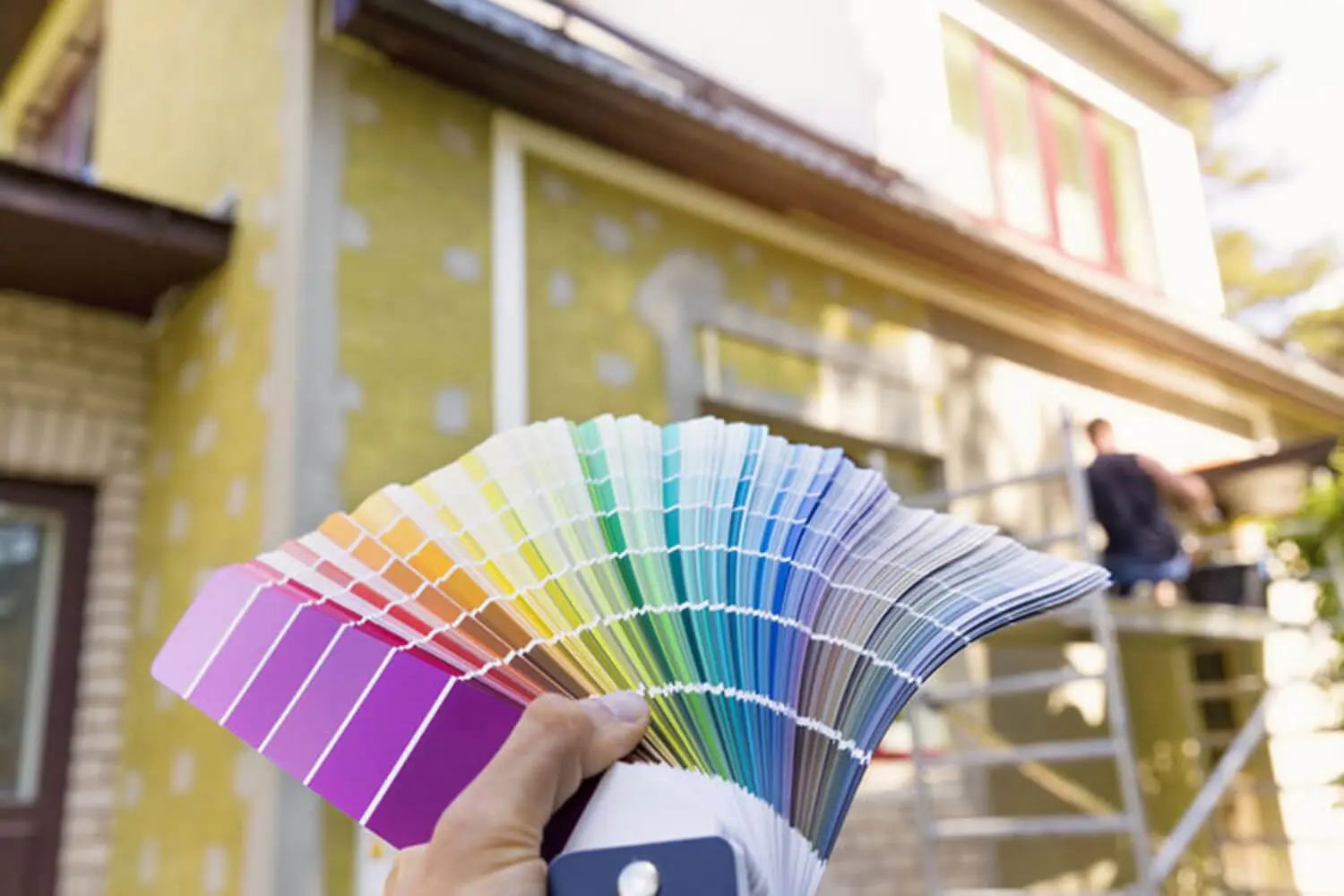 Homeowner holding paint color swatches in front of a home exterior being painted by a professional