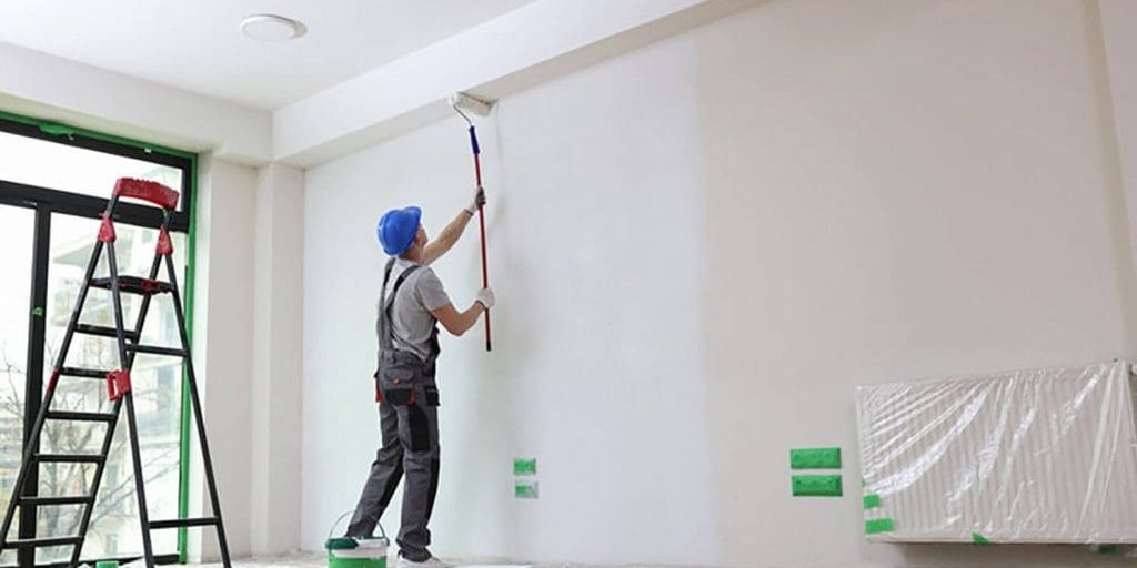 Professional painter rolling white paint on an interior wall with drop cloths and ladder in place