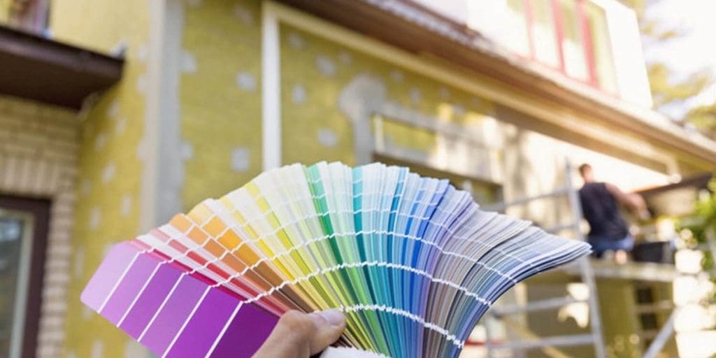 Homeowner holding paint color swatches in front of a home exterior being painted by a professional